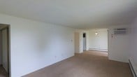 This single-floor space presents an open and functional layout with a generously sized living area finished with wall-to-wall carpet flooring in a neutral tan tone. The walls are painted crisp white throughout, contributing to a clean, bright atmosphere that makes the space feel open and airy. The ceiling features a textured finish and sits at a standard height, maintaining a comfortable sense of enclosure without feeling confined.

A baseboard heater runs along the far wall, indicating an electric heating system well-suited for year-round climate control. A doorway opening from the main living area leads toward a separate kitchen space, where a full-sized refrigerator is visible, hinting at a practical kitchen layout. A pendant light fixture hangs in the transitional area near the kitchen entrance, providing focused overhead illumination.

A wall-mounted light switch and a single electrical outlet are visible on the main living area wall, reflecting a straightforward and utilitarian design. The open flow between the living area and kitchen promotes ease of movement and versatility in how the space can be arranged and utilized.