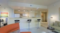 This single-story space showcases a thoughtfully designed open-concept layout that integrates the kitchen and living areas into one cohesive environment. The kitchen is equipped with white cabinetry, a full-size refrigerator, and standard appliances, all illuminated by a track lighting system mounted on the ceiling that provides directed, functional light across the workspace. A peninsula-style breakfast bar extends from the kitchen, offering casual seating with three contemporary adjustable bar stools featuring sleek chrome bases and black seats. The countertops are light-toned, complementing the neutral cream and white color scheme that runs throughout the space. The living area transitions naturally from the kitchen, with light-colored carpeting underfoot and an orange accent chair visible in the foreground, adding a pop of color to the otherwise subdued palette. A hallway visible in the background suggests additional rooms beyond the main open area, with recessed lighting guiding the way. The overall ambiance is clean, modern, and functional, making efficient use of the available floor space while maintaining an uncluttered, airy feel.