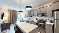 This thoughtfully designed one-bedroom, one-bathroom unit offers an efficient and functional single-floor layout with a cohesive modern aesthetic. Light wood-tone flooring flows throughout, complementing the bright white walls and contemporary furnishings that give the space an open, airy feel.

The open-plan kitchen and living area serves as the heart of the unit. The kitchen features taupe-toned cabinetry with frosted glass upper panels, stainless steel appliances including a range, microwave, dishwasher, and refrigerator, and a white quartz countertop island with bar seating beneath striking teal pendant lights. The living area accommodates a comfortable sofa, media console with a wall-mounted flat-screen TV, and direct access to a private balcony with outdoor views, allowing natural light to fill the space through floor-length drapes.

The dedicated entrance includes a built-in closet with open shelving for added storage. The bedroom comfortably fits a king-sized bed with a natural wood headboard, flanked by matching nightstands and soft ambient lighting. Built-in open shelving in the bathroom accompanies a wood-toned vanity with white countertop and a lighted mirror, rounding out this well-appointed unit.