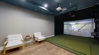 Located in Orlando, FL, this dedicated indoor golf simulator room offers a well-appointed, single-room facility designed for an immersive golf experience. The space is divided into two functional zones: a hitting bay and a spectator/lounge area, all unified by warm hardwood-style flooring and neutral gray walls.

The simulator bay dominates one end of the room, featuring a large artificial turf hitting mat that provides a realistic feel underfoot. A ceiling-mounted projector delivers vivid imagery onto a full-sized projection screen, bringing virtual golf courses to life. Overhead, a ceiling-mounted sensor bar works in tandem with the projector to accurately track swings. The bay wall is finished in a deep matte black, minimizing distraction and enhancing screen visibility. Additional club and equipment storage is accessible along the side wall.

The lounge area provides a comfortable space for spectators, furnished with cushioned armchairs with gold-tone frames and a glass side table. A wall-mounted flat-screen TV adds an additional entertainment option. A wooden storage cabinet completes the seating zone. Recessed lighting and a teal-painted ceiling add a polished, modern aesthetic throughout. The room is accessed through a stylish wood-and-frosted-glass door, contributing to the space's clean, contemporary design.
