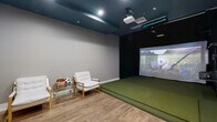 Located in Orlando, FL, this dedicated indoor golf simulator room offers a well-appointed, single-room facility designed for an immersive golf experience. The space is divided into two functional zones: a hitting bay and a spectator/lounge area, all unified by warm hardwood-style flooring and neutral gray walls.

The simulator bay dominates one end of the room, featuring a large artificial turf hitting mat that provides a realistic feel underfoot. A ceiling-mounted projector delivers vivid imagery onto a full-sized projection screen, bringing virtual golf courses to life. Overhead, a ceiling-mounted sensor bar works in tandem with the projector to accurately track swings. The bay wall is finished in a deep matte black, minimizing distraction and enhancing screen visibility. Additional club and equipment storage is accessible along the side wall.

The lounge area provides a comfortable space for spectators, furnished with cushioned armchairs with gold-tone frames and a glass side table. A wall-mounted flat-screen TV adds an additional entertainment option. A wooden storage cabinet completes the seating zone. Recessed lighting and a teal-painted ceiling add a polished, modern aesthetic throughout. The room is accessed through a stylish wood-and-frosted-glass door, contributing to the space's clean, contemporary design.