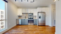 This single-floor unit offers a thoughtfully designed layout that balances modern aesthetics with functional living. The kitchen is a standout feature, showcasing a two-tone cabinet design with slate-blue lower cabinets and crisp white upper cabinets, including open shelving for display or easy-access storage. A textured tile backsplash adds visual interest, while a full suite of stainless steel appliances — including a French door refrigerator with in-door water and ice, an electric range, over-the-range microwave, and dishwasher — rounds out the space. A matte black faucet over a single-basin sink adds a contemporary finishing touch. Rich, warm-toned hardwood floors extend throughout the main living and sleeping areas, lending a sense of cohesion and warmth to the space. The bedroom benefits from large floor-to-ceiling windows that flood the room with natural light and provide views of the surrounding exterior, along with a ceiling fan for added comfort. The bathroom is finished in a clean, neutral palette featuring marble-look tile flooring, white subway-tiled tub surround, and white cabinetry accented with matte black hardware. Track lighting in the kitchen and an efficient, open floor plan make this unit both practical and visually appealing.