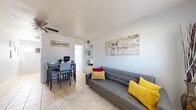 This single-floor residence offers a practical and welcoming layout with a cohesive design carried throughout by light tile flooring and soft, neutral wall tones. The open living and dining area serves as the heart of the home, featuring a tufted gray sofa accented with vibrant yellow and red pillows, a ceiling fan overhead, and a large framed abstract artwork that adds a creative focal point. A dining table with seating for four sits adjacent to the living space, making the area well-suited for both relaxing and entertaining. Natural light filters through blinds, keeping the space bright and airy. The galley-style kitchen is efficiently arranged with white cabinetry, a range, and countertop space, complemented by a window that draws in additional natural light. The two bedrooms are comfortably sized, each equipped with a ceiling fan and decorated with wall art that adds personality. One bedroom features a full-sized bed with a classic blue floral motif, while the second bedroom offers a twin bed setup with light blue walls and a window dressed with teal curtains. The bathroom is accessible from the hallway and includes a shower-tub combination with a decorative curtain and tile flooring throughout. Overall, this residence provides a functional, cheerful, and well-appointed living environment.