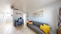 This single-floor residence offers a practical and welcoming layout with a cohesive design carried throughout by light tile flooring and soft, neutral wall tones. The open living and dining area serves as the heart of the home, featuring a tufted gray sofa accented with vibrant yellow and red pillows, a ceiling fan overhead, and a large framed abstract artwork that adds a creative focal point. A dining table with seating for four sits adjacent to the living space, making the area well-suited for both relaxing and entertaining. Natural light filters through blinds, keeping the space bright and airy. The galley-style kitchen is efficiently arranged with white cabinetry, a range, and countertop space, complemented by a window that draws in additional natural light. The two bedrooms are comfortably sized, each equipped with a ceiling fan and decorated with wall art that adds personality. One bedroom features a full-sized bed with a classic blue floral motif, while the second bedroom offers a twin bed setup with light blue walls and a window dressed with teal curtains. The bathroom is accessible from the hallway and includes a shower-tub combination with a decorative curtain and tile flooring throughout. Overall, this residence provides a functional, cheerful, and well-appointed living environment.