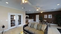 This single-story home showcases an open-concept floor plan that seamlessly connects the living room, dining area, and kitchen. The living room serves as the central gathering space, featuring light beige tile flooring, crown molding, and two ceiling fans with decorative light fixtures that provide both comfort and illumination. Recessed lighting throughout the ceiling adds a modern touch to the overall design.

The living room is anchored by a dark leather sofa adorned with striped accent pillows, creating a comfortable and inviting atmosphere. A standout feature is the set of white French doors that open to an adjacent room, allowing natural light to filter into the space. The walls are painted in a rich, deep brown tone that adds warmth and depth to the interior.

The kitchen is visible from the living area and features white cabinetry, a breakfast bar with a light countertop, and standard appliances. The dining area, positioned nearby, includes a dark table and chairs alongside a decorative potted plant. The cohesive layout and warm color palette create a functional and welcoming living environment throughout the home.