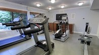This fitness center is a dedicated single-floor workout space designed to support a well-rounded cardio routine. The room is outfitted with multiple commercial-grade cardio machines, including treadmills and an elliptical trainer from True Fitness, providing users with a variety of exercise options. A wall-mounted flat-screen television is positioned prominently, allowing exercisers to stay entertained during their workouts. The space features light gray walls accented with clean white crown molding and baseboards, lending a polished, well-maintained appearance. Flush-mounted ceiling lights provide consistent, even illumination throughout the room. A window along one wall allows natural light to filter in, offering a pleasant view of outdoor greenery and contributing to an open, airy atmosphere. Warm wood-toned cabinetry beneath the television provides practical storage and adds a touch of warmth to the otherwise neutral palette. The flooring is a smooth, durable surface suited for fitness use. Overall, the fitness center is thoughtfully arranged to maximize usable space while maintaining a clean, comfortable environment conducive to exercise.