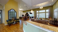 Located at 9200 Bunsen Park Dr in Louisville, KY, this single-floor community clubhouse offers a well-designed, open-concept layout that seamlessly integrates multiple functional areas into one cohesive space. The entryway greets visitors with a curved granite-topped reception counter and a striking arched mirror, setting an inviting tone throughout.

The expansive lounge area features soaring ceilings, olive-green accent walls, warm hardwood flooring, and large windows that flood the space with natural light. Comfortable seating arrangements—including a tufted sofa, armchairs, and a glass-top coffee table—are complemented by oversized abstract artwork, creating a relaxed yet refined ambiance.

A fully equipped kitchen anchors one end of the space, outfitted with dark cabinetry, granite countertops, stainless steel appliances, and a bar-height island. Dual wall-mounted flat-screen TVs add a media-forward element to the kitchen area.

Multiple dining configurations accommodate a variety of gatherings. A formal dining nook features a dark wood table with upholstered tufted chairs beneath a chandelier, while a casual high-top bar area provides a more relaxed setting with round tables and counter-height seating. Each dining area benefits from large windows and wall-mounted screens. The overall design balances comfort, functionality, and style throughout.