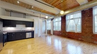 This loft-style studio with two bathrooms showcases a compelling blend of industrial character and modern finishes throughout its open, single-floor layout. The main living area commands attention with soaring wood-plank ceilings supported by white-painted beams, exposed silver ductwork running overhead, and gleaming light hardwood floors that extend throughout the space. A row of large multi-pane windows set within original exposed brick walls lines one side of the room, drawing in abundant natural light and offering views of the surrounding area. Pendant globe fixtures complement recessed and track lighting for versatile illumination.

The kitchen is well-appointed with dark navy cabinetry, granite countertops, and a full suite of stainless steel appliances including a refrigerator, electric range, and dishwasher. Generous cabinet storage reaches toward the ceiling for added functionality.

Two bathrooms provide distinct experiences: one features dark slate-style tile floors, a vanity with dark countertop, and a walk-in shower with floor-to-ceiling tile, while the second includes a tub-shower combination with matching dark tile surround. Both bathrooms maintain a cohesive, modern aesthetic that complements the industrial character of the overall space.