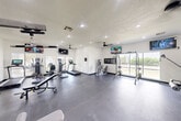 Located at 1512 Hollowhill Drive in Bryan, Texas, this fitness center offers a clean, well-appointed exercise room designed to support a comprehensive workout experience. The space features durable rubber flooring throughout, providing a safe and functional surface for all types of exercise. Recessed lighting and ceiling fans ensure the room remains bright and comfortable during any workout session.

The cardio zone is equipped with treadmills, an elliptical trainer, and a stationary bike — all from the True Fitness brand — positioned alongside a large wall-mounted mirror that enhances the sense of space and allows users to monitor their form. Multiple flat-screen televisions are mounted throughout the room, offering entertainment during workouts.

The strength training area includes a cable crossover machine, a leg extension and curl machine, an adjustable weight bench, a lat pulldown machine, and a full rack of dumbbells organized on a tiered stand. A power rack with cable attachments rounds out the free weights section, providing versatility for a wide range of exercises.

Large windows along one wall invite natural light into the space and offer views of the surrounding greenery. The room opens through glass-panel doors to an adjacent area, and a hand sanitizer station reflects a commitment to cleanliness. This fitness center is a well-rounded, functional facility suited for residents seeking convenient on-site exercise amenities.