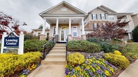 The Islands of Fox Chase Leasing Center serves as the welcoming gateway to this residential community. The building's exterior presents a charming, traditional architectural style, highlighted by prominent white columns framing a covered front porch, a graceful arched entryway, and a combination of beige lap siding and natural stone accents that lend the structure both warmth and character. A set of concrete steps with black iron railings leads visitors up to the front entrance, which is framed by well-maintained landscaping featuring vibrant seasonal flowers in yellow and purple, neatly trimmed hedges, and lush greenery that adds a colorful and inviting curb appeal. The surrounding community grounds are equally well-kept, with wide, smooth roadways lined by continuous sidewalks, ample parking, and mature trees displaying rich autumn foliage that enhances the overall ambiance of the neighborhood. Multi-story residential buildings are visible throughout the community, all sharing a cohesive aesthetic with matching siding and stone detailing. Rolling landscaped hills with natural woodland backdrops provide a scenic setting. The leasing center's single-floor layout ensures easy accessibility, while its prominent signage and well-lit entrance make it straightforward to locate within the community.