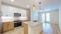 This thoughtfully designed single-floor residence showcases a contemporary aesthetic with clean lines and a bright, airy atmosphere throughout. The open-concept layout seamlessly connects the kitchen and living area, creating a cohesive and functional living space.

The kitchen is a standout feature, anchored by a large central island topped with speckled granite countertops and equipped with a professional-style pull-down faucet and undermount sink. A mix of white upper cabinetry and warm natural wood lower cabinets adds visual depth, complemented by a classic white tile backsplash. Stainless steel appliances — including a refrigerator with ice dispenser, electric range, and built-in microwave — complete the modern kitchen. Amber glass pendant lights add warmth and character overhead.

Light wood-style flooring flows through the main living areas, enhancing the sense of openness and continuity. The living area benefits from natural light through large windows and a glass door, with a ceiling fan providing additional comfort.

The bedrooms feature plush carpeting and generous wall space, offering a soft, quiet retreat. The bathroom is well-appointed with white shaker-style cabinetry, a single vanity with a light countertop, a large framed mirror, and overhead storage. Light wood flooring continues into the bathroom, maintaining a cohesive design throughout the unit.