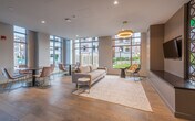The amenity level at 1 Chestnut Place is a thoughtfully designed, single-floor community space that blends comfort, functionality, and contemporary style. The layout is divided into distinct zones, each serving a specific purpose while maintaining a cohesive aesthetic throughout.

The main lounge area is anchored by a plush neutral sofa atop a woven area rug, complemented by accent chairs and a small side table. Floor-to-ceiling windows wrap the perimeter, flooding the space with natural light and offering direct views of a landscaped outdoor courtyard. A statement pendant chandelier with a warm gold finish adds an elegant focal point overhead, while light hardwood flooring runs throughout, lending warmth to the modern design.

An adjacent lounge and bar area shifts to a more refined atmosphere, featuring white upholstered sofas, a gold and marble-top coffee table, and a built-in wet bar with a beverage cooler. Dark, iridescent tile creates a dramatic backsplash, while pendant lights with brass accents and a bold piece of wall art add personality. Bar-height seating lines a countertop along a textured accent wall.

A quiet work nook with a built-in desk, wall-mounted task lighting, and accessible power outlets offers a focused workspace. Additional corridors connect the zones seamlessly, finished with clean white walls and recessed lighting for a polished, move-in-ready feel.
