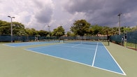This outdoor tennis facility features multiple hard courts finished in a classic blue and green color scheme, with crisp white boundary lines clearly marking each playing area. The courts are constructed with a hard, even surface well-suited for competitive and recreational play alike. Each court is equipped with a regulation net, and the complex is fully enclosed by chain-link fencing that defines the perimeter and helps contain play within each court.

Tall floodlight poles are positioned throughout the facility, indicating the courts are equipped for evening play. The surrounding landscape is lush, with mature trees and tropical foliage lining the boundary of the complex, providing a natural visual buffer and partial shade along the edges. The greenery creates an appealing backdrop and contributes to a pleasant outdoor atmosphere.

The facility appears to accommodate multiple simultaneous matches, with clearly delineated court spacing. Paved walkways and access points allow for easy movement between courts. The overall setting suggests a well-maintained recreational or community sports complex, designed to support consistent use by players of varying skill levels in an open-air environment.