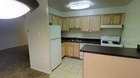 This single-floor space centers around a practical and neatly designed kitchen that serves as a key feature of the layout. The kitchen is outfitted with light natural wood cabinetry that extends to the upper and lower sections of the walls, providing generous storage space. Dark laminate countertops offer a contrasting aesthetic, complementing the light-toned cabinetry and creating a clean, classic look. White appliances, including a full-size refrigerator, a gas range, and a dishwasher, are seamlessly integrated into the layout. A stainless steel double sink is positioned beneath upper cabinets, offering a practical workstation. The kitchen floor is finished with light-colored square tiles, which add brightness to the space and are easy to maintain. A peninsula or partial counter extends outward, providing additional prep space or the potential for casual seating. Overhead, a flush-mount circular ceiling light provides even, functional illumination throughout the kitchen. The space transitions naturally from the tiled kitchen into what appears to be a carpeted adjoining area, suggesting a connected and cohesive floor plan designed for comfortable, everyday living.