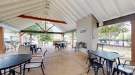 Located in Lakeland, Tennessee, this generously sized grilling pavilion offers a welcoming and functional outdoor gathering space defined by its striking architectural design. The centerpiece of the structure is a dramatic vaulted ceiling with exposed white-painted wooden beams and rafters, supported by bold cross-braced timber trusses that lend the space a classic, open-air pavilion character. Ceiling fans are mounted along the ridge to ensure comfortable airflow throughout.

The aggregate concrete flooring spans the entire covered area, providing a durable and easy-to-maintain surface. Multiple seating arrangements, including metal bistro-style tables and chairs as well as round patio tables, are distributed throughout, comfortably accommodating groups of various sizes.

A built-in outdoor kitchen station with a stainless steel grill and brick surround serves as a functional cooking hub, complemented by a full-height white brick fireplace that adds warmth and ambiance for cooler evenings. The open sides of the pavilion frame views of the surrounding mature trees and landscaped grounds, with a fenced swimming pool area visible nearby. The pavilion's open design allows abundant natural light to fill the space, creating a bright and inviting atmosphere ideal for outdoor entertaining and community gatherings.