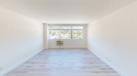 This thoughtfully renovated single-floor residence offers a clean, modern aesthetic throughout, defined by crisp white walls, light hardwood flooring, and an abundance of natural light. The open living area is a standout feature, anchored by large picture windows that frame sweeping views of the surrounding hillside and neighborhood below, creating a bright and airy atmosphere. A wall-unit air conditioner ensures year-round comfort in the main living space.

The kitchen is sleek and highly functional, featuring high-gloss white cabinetry extending to the ceiling, light quartz countertops, recessed lighting, and a full suite of stainless steel appliances including a refrigerator, electric range, built-in microwave, and dishwasher. The layout provides ample counter space and storage, making it well-suited for everyday cooking.

The bedrooms are generously proportioned, with the same light hardwood flooring and neutral white walls that carry throughout the unit. Multiple windows in the bedroom areas continue the theme of natural light and hillside views, with one room featuring a mirrored closet. The bathroom is neatly appointed with a white vanity, tile flooring, recessed lighting, a large mirror, and a glass-enclosed shower and tub combination. Overall, the space presents a cohesive, contemporary feel with clean finishes throughout.