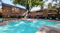This outdoor pool area serves as a central amenity within a multi-story brick and wood-sided residential community. The centerpiece is a generously sized rectangular swimming pool with crystal-clear blue water, accessible via stainless steel entry rails. Adjacent to the pool sits a spa, offering a dedicated relaxation option just steps from the main pool. The expansive concrete sundeck surrounds the pool on multiple sides and is furnished with a collection of lounge chairs and patio seating, providing ample space for sunbathing and outdoor leisure. Shade umbrellas are positioned throughout the deck, offering respite from direct sunlight. The area is framed by mature trees whose broad canopies cast natural shade over portions of the deck, enhancing the overall comfort of the outdoor space. Lush landscaping, including shrubs and ornamental plantings, lines the perimeter, contributing to a well-maintained and inviting atmosphere. A dedicated pool house structure with rustic wood-framed glass doors provides access to the pool area and includes a covered porch with additional seating. The building facades of the surrounding multi-story structures, featuring brick accents and private balconies, create an enclosed, community-oriented setting. A black iron perimeter fence defines the pool area while maintaining open sightlines to the surrounding grounds.
