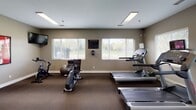 This fitness center offers a comprehensive and well-organized workout environment spread across a single floor. The space is finished with durable dark carpeted flooring and neutral taupe-toned walls, creating a clean and focused atmosphere conducive to exercise.

The cardio zone features multiple treadmills, elliptical trainers, and upright bikes, each equipped with individual display screens for entertainment during workouts. Large windows fitted with horizontal blinds line the walls, inviting natural light and providing views of the surrounding greenery. A full-length wall mirror in the cardio area visually expands the space and allows users to monitor their form.

The strength training area is anchored by a professional-grade cable machine system capable of a wide variety of resistance exercises. A spin bike rounds out the equipment offerings in this zone, alongside a wall-mounted flat-screen television. Sports-themed artwork adorns the walls throughout, adding energy and personality to the environment.

Flush-mounted ceiling light fixtures provide consistent, bright illumination throughout the space. Structural columns are integrated thoughtfully into the layout without impeding the flow of the room. A stability ball and additional accessories are also available, supporting a well-rounded fitness routine. The overall design strikes a balance between functionality and comfort, making it an inviting space for daily exercise.