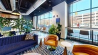 Located at 104 Washington St in downtown Dover, New Hampshire, this single-floor commercial coworking space offers a thoughtfully designed environment that balances productivity with comfort. The open-plan layout is anchored by polished concrete floors and an exposed black ceiling with industrial-style ductwork, creating a modern, urban aesthetic throughout.

The entrance corridor features a warm wood-slat ceiling, bold navy accent walls, and a built-in stepped bench seating area with decorative pillows — an inviting welcome for members and guests. Floor-to-ceiling glass partitions line the hallway, providing visual connectivity between zones while maintaining a sense of structure.

The main coworking area offers rows of shared workstations topped with acoustic privacy panels and illuminated by stylish pendant lights. Lush indoor plants are strategically placed throughout, adding a natural, refreshing contrast to the industrial design elements. A vibrant lounge zone near the large street-facing windows features an eclectic mix of seating — navy sofas, leather club chairs, and high-top tables — all bathed in abundant natural light. A dedicated dining/breakout area includes upholstered booth seating in deep green, pendant lighting, and curated wall art, providing a relaxed setting for informal meetings or breaks.