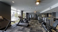 This fitness center offers a comprehensive workout environment designed with both functionality and comfort in mind. Floor-to-ceiling windows line one side of the space, flooding the interior with natural light and providing open views of the surrounding area, creating an energizing atmosphere for exercise. The patterned carpet flooring adds texture and warmth while defining the workout zones throughout the room.

The cardio section is prominently positioned near the windows and features multiple treadmills, elliptical machines, and stationary bikes, allowing users to exercise while enjoying the outdoor views. Ceiling fans are installed overhead, promoting airflow and comfort during workouts. Recessed lighting complements the natural light, ensuring the space remains well-lit throughout the day.

Toward the rear, a dedicated strength-training area is outfitted with weight machines and resistance equipment. Large mirrors line the back wall, visually expanding the space and allowing users to monitor their form. The dark gray accent walls contrast with the bright, window-lit front section, giving the fitness center a modern, polished aesthetic. Overall, the space is thoughtfully designed to accommodate a full range of fitness activities.