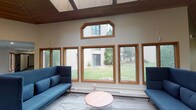 This single-floor space presents a welcoming communal lounge defined by distinctive architectural character and thoughtful design. The most striking feature is the vaulted wood-paneled ceiling, finished in warm tones that lend the interior a natural, inviting quality. A suspended rectangular lighting fixture with recessed downlights anchors the central area, complementing the overall brightness of the space.

Large wood-framed windows line the walls, including a prominent angled transom window positioned above a bank of wider panes, allowing generous natural light to pour in throughout the day. These windows frame views of a green lawn and surrounding outdoor scenery, creating a strong visual connection to the exterior environment.

The lounge is furnished with blue upholstered sofas and accent chairs arranged around a round, blush-toned coffee table, forming a comfortable seating area suited to conversation or relaxation. Patterned carpeting in neutral tones covers the floor, adding texture underfoot. A service counter or reception-style station is visible toward the back of the space, alongside a beverage vending machine, suggesting this area serves as a common gathering hub. The overall ambiance is open, airy, and casually functional.