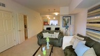 This three-floor residence is thoughtfully designed to provide both comfort and practicality across its multiple levels. The main living area serves as a welcoming focal point, featuring plush carpeting underfoot and warm neutral-toned walls that create a relaxed, cozy atmosphere. The space comfortably accommodates generous seating arrangements, highlighted by a deep-cushioned sofa set paired with a glass-top coffee table, adding a contemporary touch to the room.

A raised breakfast bar visually separates the living area from the kitchen space beyond, where pendant lighting hangs above, casting a warm glow throughout the open layout. The kitchen area features bar-style seating along the counter, making it ideal for casual dining or entertaining. A window above the kitchen counter allows natural light to filter into the space.

A doorway visible from the living area provides access to an adjacent room, likely a bathroom or utility space, further enhancing the functional layout of this level. The staircase, visible in the background, connects the floors, offering easy access to the upper and lower levels of this multi-story residence. Overall, the space presents a warm, well-organized home environment.