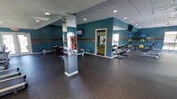 This fitness center offers a generous, open workout space designed to accommodate a variety of exercise routines. The room features durable rubber flooring in a dark speckled finish, providing both safety and comfort underfoot. Teal-painted walls accented with a horizontal stripe add a dynamic, energetic aesthetic to the space.

A mirrored column positioned in the center of the room enhances the sense of space while providing reflective surfaces useful during workouts. Recessed lighting in the drop ceiling ensures the area is well-lit throughout. Mounted flat-screen televisions are positioned for easy viewing from multiple workout stations.

The strength training area is equipped with a dumbbell rack stocked with a full range of weights, along with additional free weights and accessories. Cardio equipment, including rowing machines, is arranged along the perimeter. A vending machine is available for convenience. A doorway at the rear of the space leads to an additional area, with windows offering natural light. Wall-mounted speakers complete the facility, providing an engaging workout environment.