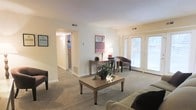 This single-story residence features a welcoming living area defined by neutral-toned walls and wall-to-wall carpeting that lends a warm, comfortable feel throughout. The space is anchored by a generously sized living room that benefits from an abundance of natural light, courtesy of a set of French doors that open to an outdoor area, creating a seamless connection between interior and exterior. The doors are framed by blinds, offering flexible light control while maintaining a clean, understated aesthetic.

The room accommodates a practical and classic furniture arrangement, including a central coffee table, accent seating, and a console table dressed with a table lamp and decorative artwork above it, adding a touch of personality to the space. A visible hallway leads to additional rooms within the unit, including what appears to be a well-lit bathroom or utility area. Ceiling vents are distributed throughout, indicating a central HVAC system for year-round climate control. The overall ambiance is calm and functional, with a traditional design sensibility and a layout that maximizes usability across its single floor.