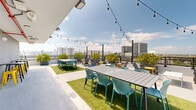 Located at 1642 NW 16th St in Miami, FL, this rooftop terrace offers a vibrant and well-appointed outdoor gathering space with panoramic views of the Miami skyline. The terrace is designed to accommodate a variety of social settings, featuring multiple dining tables with teal lattice chairs arranged atop patches of artificial turf, creating a lush contrast against the smooth concrete flooring. String lights draped overhead add a warm, festive ambiance suitable for both daytime and evening use. A dedicated lounge area outfitted with Adirondack-style chairs and side tables provides a more relaxed setting for guests to take in the expansive city views. The space also includes a bar-height seating zone with bold yellow stools, ideal for casual gatherings. A built-in stainless steel grill station positioned along the perimeter wall makes the terrace fully equipped for outdoor cooking and entertainment. Lush potted shrubs line the railings, adding greenery and a sense of privacy. Floor-to-ceiling sliding glass doors connect the terrace to an interior space, allowing for seamless indoor-outdoor flow. The sturdy metal railing system borders the terrace on all sides, ensuring safety while preserving unobstructed sightlines to the surrounding Miami cityscape and open sky.