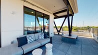 Located at 240 Grant St SE in Atlanta's Grant Park neighborhood, this contemporary two-floor amenity space combines sophisticated interior design with striking outdoor living areas. The interior lounge is appointed with plush navy leather seating arranged around a walnut coffee table, anchored by a wall-mounted flat-screen television recessed into a clean white surround. Large floor-to-ceiling windows flood the space with natural light while framing views of the surrounding Atlanta skyline. Adjacent to the lounge, a stylish wet bar area features a bold tiled backsplash, geometric pendant lighting, under-counter cabinetry, and a generous island with woven bar stools — ideal for entertaining. Slate-toned tile flooring runs throughout, lending a cohesive, modern aesthetic. Expansive glass doors open directly onto the rooftop terrace, where bold black structural steel cross-bracing adds an industrial architectural accent. The terrace offers ample open-air space with built-in bench seating, a covered lounge area with comfortable outdoor sofas, and panoramic views of the Atlanta cityscape and surrounding greenery. A dedicated outdoor dining counter with bar seating extends the entertaining space seamlessly between indoors and out, making this a versatile and well-appointed urban retreat.