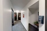 Located at 8713 Drumlin Heights Dr in Baldwinsville, NY, this single-floor common area serves as a functional shared space within a residential building. The layout comprises two hallways and a dedicated laundry room, efficiently organized to serve the building's residents.

The hallways feature dark tile flooring with a subtle patterned texture, crisp white walls, and flush ceiling lighting that keeps the corridors well-lit. Bold dark-framed unit entry doors line the hallways, each clearly numbered for easy identification. A fire extinguisher is visibly mounted on the wall, reflecting attention to building safety standards. A small decorative accent table with a potted plant adds a welcoming touch to one of the corridor alcoves.

At the end of the hallway, a windowed exit door provides clear egress from the building. The laundry room houses Whirlpool top-loading washer and dryer units and includes an electrical panel, offering residents a convenient on-site laundering option. The room is finished with the same clean white walls found throughout the common areas, maintaining a consistent and tidy aesthetic. Overall, this shared space is practical and well-maintained, providing essential amenities and safe, easy navigation for building residents.