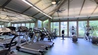 This single-story fitness center is designed to provide a bright and motivating workout environment. The space is defined by its striking vaulted ceiling with exposed structural beams and white-painted wood paneling, lending an open and airy architectural character. A ceiling fan is centrally mounted, helping to maintain comfortable airflow throughout the room.

Expansive windows wrap around multiple walls, flooding the interior with natural light and offering unobstructed views of surrounding greenery and neighboring structures. This connection to the outdoors contributes to a calm, refreshing atmosphere that distinguishes the space from typical enclosed gym settings.

The flooring consists of durable, rubberized tile in a neutral tone, well-suited for fitness use. The facility is equipped with a variety of cardio machines, including treadmills, elliptical trainers, stationary bikes, and stair-stepping equipment — offering a comprehensive range of aerobic exercise options. Equipment brands such as Cybex are notably present, indicating a commitment to quality.

Mirrors along one wall enhance the sense of space and allow users to monitor form. The overall ambiance is welcoming, clean, and functional, balancing practical fitness amenities with an architecturally appealing, light-filled setting.