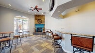 This single-story community amenity center is thoughtfully designed to offer residents a variety of gathering and recreational spaces, all unified by a warm color palette of teal accents, neutral walls, and rich wood tones. The central lounge area makes an immediate impression with its striking floor-to-ceiling stacked stone fireplace, accented by a teal mantle surround. High-top tables with wrought iron bar stools are arranged throughout, complemented by patterned carpet tile flooring and ceiling fans that enhance the comfortable, social atmosphere. A granite-topped bar counter with additional seating anchors one side of the room, while a wall-mounted television adds to the entertainment appeal. Adjacent to the lounge, the kitchenette is outfitted with rich wood cabinetry, granite countertops, slate tile flooring, and full-sized appliances including a built-in refrigerator and microwave. The dedicated screening room features rows of cushioned theater-style seating facing a large flat-screen television set within a handsome wood entertainment wall, with high clerestory windows allowing subtle natural light. The expansive fitness center is comprehensively equipped with strength machines, treadmills, ellipticals, stationary bikes, and stability balls, all set within a bright space with arched windows, mirrored walls, and direct exterior access.
