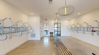 Located at 1181 Manhattan Blvd in Dayton, Kentucky, this dedicated bike room is a thoughtfully designed amenity space that combines functionality with modern aesthetic appeal. The single-floor layout offers a generous, open feel highlighted by polished concrete floors that reflect natural light streaming in through multiple windows flanking the exterior entry door.

Wall-mounted bike racks line multiple walls, providing organized, secure storage for numerous bicycles. A standout feature is the dedicated bike wash station, tiled in crisp white subway tile with a floor drain and hose hookup, making maintenance convenient and mess-free. A red bike repair stand is also present, offering a practical space for on-site tune-ups.

The space is anchored by a sleek, slatted metal bench at its center, ideal for suiting up or winding down. Overhead, sculptural wire pendant light fixtures add an unexpected design element, elevating the room beyond purely utilitarian use. Recessed lighting complements the natural light, keeping the space bright and inviting. A black steel-framed glass interior door provides a stylish visual connection to adjacent areas of the building.

The clean white walls, modern fixtures, and organized layout make this bike room a practical yet refined amenity for cycling enthusiasts.