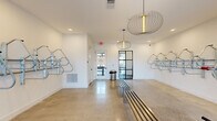 Located at 1181 Manhattan Blvd in Dayton, Kentucky, this dedicated bike room is a thoughtfully designed amenity space that combines functionality with modern aesthetic appeal. The single-floor layout offers a generous, open feel highlighted by polished concrete floors that reflect natural light streaming in through multiple windows flanking the exterior entry door.

Wall-mounted bike racks line multiple walls, providing organized, secure storage for numerous bicycles. A standout feature is the dedicated bike wash station, tiled in crisp white subway tile with a floor drain and hose hookup, making maintenance convenient and mess-free. A red bike repair stand is also present, offering a practical space for on-site tune-ups.

The space is anchored by a sleek, slatted metal bench at its center, ideal for suiting up or winding down. Overhead, sculptural wire pendant light fixtures add an unexpected design element, elevating the room beyond purely utilitarian use. Recessed lighting complements the natural light, keeping the space bright and inviting. A black steel-framed glass interior door provides a stylish visual connection to adjacent areas of the building.

The clean white walls, modern fixtures, and organized layout make this bike room a practical yet refined amenity for cycling enthusiasts.