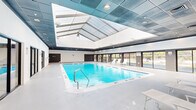Located at 275 Research Pkwy in Meriden, Connecticut, this single-floor fitness and aquatic facility offers a comprehensive wellness environment designed for both recreation and exercise. The centerpiece of the space is a generously sized indoor swimming pool, bathed in natural light streaming through a striking vaulted skylight overhead. Large floor-to-ceiling windows line the pool area, providing views of the surrounding landscape and enhancing the open, airy atmosphere. White lounge chairs are positioned poolside for relaxation, and a separate hot tub area adds to the aquatic amenities. Adjacent to the pool, a dedicated yoga studio features warm wood-look flooring, full-length mirrored walls, and a clean white interior — ideal for group classes or individual practice. The main fitness floor is expansive and well-equipped, featuring treadmills, strength training machines, and free weights set on durable speckled flooring. Bold accent walls in magenta energize the workout area. Throughout the facility, dark tile ceilings with recessed lighting create a modern, polished aesthetic. An additional open multipurpose room offers flexible space for various fitness activities. The overall design balances functionality with comfort, creating an inviting environment for all wellness pursuits.