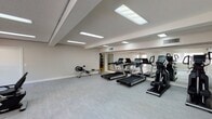 This fitness center is designed to support a well-rounded cardio workout routine within a clean, functional layout. The space is finished with light-toned vinyl plank flooring that lends a modern, low-maintenance aesthetic throughout. A textured ceiling hosts multiple recessed fluorescent panel lights, ensuring the room is evenly and brightly illuminated. Natural light supplements the artificial lighting through windows along one wall, contributing to an open and energizing atmosphere.

The equipment selection includes multiple treadmills, an elliptical trainer, a rowing machine, a recumbent bike, and an upright stationary bike, providing diverse options for cardiovascular training. A rack of free weights is visible near a full-length mirror wall, which spans one side of the room and visually expands the space while allowing users to monitor their form during exercise. The mirrored wall also reflects natural light and the surrounding equipment, enhancing the sense of openness.

The room is accessed through a glass-panel door connecting to an adjacent common area, with a secondary exit door also present. The neutral wall tones and uncluttered floor space create a practical, distraction-free environment well-suited for focused exercise.