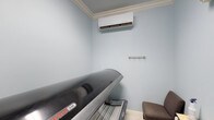 The tanning room is a compact, single-purpose space designed to provide a private and comfortable environment for indoor tanning. The room is finished with soft light blue walls and white crown molding that adds a clean, polished look to the interior. Light-toned tile flooring contributes to the bright, hygienic feel of the space.

Central to the room is a professional-grade tanning bed, model 336, which serves as the focal point of the space. A wall-mounted mini-split air conditioning unit is positioned near the ceiling, ensuring the room remains cool and comfortable during use — a particularly practical feature given the heat generated by tanning equipment. A small metal hooks rail is mounted on the wall, providing a convenient spot to hang clothing or personal items during a session. A cushioned chair is also present, offering a place to sit before or after use.

The room's thoughtful layout maximizes functionality within its modest footprint, creating a clean and well-appointed space suited for a salon, fitness facility, or wellness center.