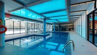 Located at 375 Rue de la Concorde in the heart of Montréal, the C-Loft amenity floor delivers a comprehensive, resort-style experience across a generously sized single level. The centerpiece is a striking indoor swimming pool bathed in vivid cyan light emanating from luminous ceiling panels, creating an immersive aquatic atmosphere. The pool area features sleek tile surrounds, lounge chairs for relaxation, large glazed walls that admit natural light, and a decorative mural that adds a vibrant, artistic touch to the space. An adjacent relaxation lounge offers additional seating with complementary décor and atmospheric accent lighting. The fully equipped fitness center is divided into distinct zones: a cardio area outfitted with treadmills, ellipticals, and rowing machines beneath an industrial-style black ceiling with exposed ductwork and bright linear lighting, and a dedicated free weights section featuring dumbbells, benches, cable machines, and full-length mirrors alongside sheer-curtained windows. A media lounge with warm carpeting, a mounted flat-screen television, and casual seating provides a comfortable retreat. Completing the layout are a functional office space and a connecting hallway that links the various zones seamlessly. The overall design balances modern industrial aesthetics with wellness-focused amenities.