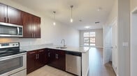 This thoughtfully designed one-bedroom, one-bathroom residence offers a cohesive and modern living experience across a single floor. The open-concept kitchen and living area serves as the heart of the home, where rich dark-cherry cabinetry pairs elegantly with granite countertops and a full suite of stainless steel appliances, including a refrigerator, electric range, microwave, and dishwasher. A peninsula with pendant lighting above creates a natural divide between the kitchen and the living area, which benefits from large windows that invite natural light and offer views of the surrounding neighborhood. Wood-style plank flooring flows seamlessly throughout the common areas and into the bedroom, providing a warm and cohesive aesthetic. The bedroom is a comfortable, well-proportioned retreat featuring a window with outdoor views and direct access to a dedicated closet outfitted with wire shelving for organized storage. The bathroom is clean and functional, featuring a dark-cherry vanity with a granite countertop, a framed mirror, and a tub-shower combination. A utility room provides additional practical storage and laundry accommodation. Neutral wall tones throughout lend the space a bright, move-in-ready feel.