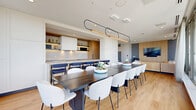 Located in Ottawa's McRae Avenue, this contemporary amenity space offers a thoughtfully designed open-concept layout spread across a single level. The interior is defined by clean lines, light hardwood flooring, and a neutral palette punctuated by warm wood accents throughout.

The kitchen is outfitted with high-end stainless steel appliances, including a built-in induction cooktop, wall oven, dishwasher, and a sleek range hood. White quartz countertops and warm wood cabinetry provide a refined, functional workspace, complemented by a bar-style counter with seating.

The dining area accommodates a large, dark wood communal table surrounded by an ample arrangement of modern chairs — ideal for group gatherings. A sculptural linear pendant light fixture anchors the space above. Open sightlines connect the dining area to a comfortable lounge furnished with a generous sectional sofa in warm amber tones, a wall-mounted television set against a slatted wood accent wall, and sheer floor-to-ceiling curtains that filter abundant natural light.

Floor-to-ceiling windows frame sweeping views of the Ottawa skyline and surrounding greenery, seamlessly connecting the interior to a spacious outdoor terrace. The terrace is equipped with multiple BBQ stations, large dining tables, and teal outdoor seating — all set against an open-sky backdrop with unobstructed panoramic views.