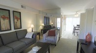 This two-floor residence offers a well-appointed living space designed for both comfort and functionality. The living room serves as the heart of the home, featuring a large mirrored wall panel that visually expands the room and enhances the sense of openness. Soft, neutral-toned carpeting runs throughout, complementing the understated color palette of the walls and furnishings.

Seating in the living area includes a charcoal sofa and a patterned armchair accented with a deep red throw pillow, arranged around a dark-toned coffee table to create an inviting conversational setting. Warm table lamps on either side of the room provide ambient lighting, adding to the cozy atmosphere.

A standout feature is the floor-to-ceiling sliding glass door at the far end of the room, fitted with vertical blinds that allow for adjustable natural light. This opening floods the space with daylight and provides access to an outdoor area, contributing to an airy, connected feel. The overall ambiance is clean and comfortable, with thoughtful design details that maximize both light and the perception of space across its two levels.