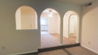 This single-floor residence showcases a distinctive architectural design centered around a series of graceful arched doorways that define and connect the main living areas. The arches, finished in a clean white with a soft gray surround, lend a classic, refined character to the interior and create a natural sense of flow throughout the space.

The carpeted living and dining areas are open and generously proportioned, offering a comfortable and welcoming atmosphere. The warm-toned carpet extends seamlessly across the main living spaces, contributing to a cozy ambiance. A ceiling fan is centrally positioned in the main living area, providing both functional air circulation and a subtle decorative element.

Beyond the arched entryways, a glimpse into the kitchen reveals white cabinetry and a well-organized layout, with natural light filtering in through a window. A separate doorway visible through the arches suggests additional rooms, providing a practical and well-divided floor plan.

The interior is finished with clean white trim, neutral-toned walls, and consistent base molding throughout, creating a cohesive and understated aesthetic. The overall design emphasizes an open, connected layout while maintaining distinct zones, making the space both functional and visually appealing.
