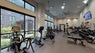 This fitness center is designed to accommodate a wide range of workout styles within a thoughtfully organized, single-floor layout. The space is characterized by soaring ceilings, large floor-to-ceiling windows along the perimeter walls that flood the interior with natural light, and views of surrounding greenery and architecture. An oversized industrial ceiling fan and pendant lighting fixtures contribute to both airflow and ambiance throughout the main workout area.

The cardio zone is well-stocked with stationary bikes, elliptical machines, treadmills, and Concept2 rowing machines, all positioned to take advantage of the natural light and outdoor views. Adjacent to this area is a dedicated strength training section featuring Rogue-brand benches, a power rack loaded with bumper plates, a full dumbbell rack, and cable machines. Full-length mirrors line the walls, enhancing the sense of space and aiding in form monitoring.

A separate functional training room offers a distinct workout environment, equipped with a ballet barre, medicine ball racks, a tire, resistance tools, and wall-mounted mirrors and television. A glass-partitioned room is also visible, suggesting a private training or stretching area. The flooring transitions between rubberized material and carpet-like surfaces, each suited to its respective training zone. Overall, the fitness center delivers a comprehensive, professionally appointed workout environment.