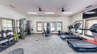 Located at 4095 Lawson Ridge Dr in Huntsville, Alabama, this fitness center offers a well-equipped, single-floor workout facility designed to support a full range of exercise routines. The space features neutral-toned walls, carpeted flooring, and ample overhead fluorescent lighting complemented by ceiling fans that maintain a comfortable atmosphere throughout.

The cardio zone is outfitted with multiple treadmills, including incline trainers, as well as elliptical machines and a stationary bike, providing a thorough selection of aerobic equipment. For strength training, the facility includes a multi-station cable machine, a leg press/extension machine, adjustable weight benches, and a dedicated free weights area stocked with a full rack of dumbbells and kettlebells in various weights. Resistance bands and exercise balls are also available for functional fitness training.

A large wall-mounted mirror expands the sense of space and allows users to monitor their form during workouts. Wall-mounted televisions add entertainment options for cardio sessions. Multiple windows fitted with blinds line the walls, allowing natural light to filter in while maintaining privacy. The fitness center connects to an adjoining common area through a clearly marked exit, making it easily accessible. Overall, the space is thoughtfully arranged to accommodate simultaneous use by multiple individuals comfortably.