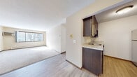 This single-floor unit offers a practical and well-organized layout designed for comfortable everyday living. The galley-style kitchen is efficiently arranged with rich dark wood cabinetry lining both sides, light stone-finish countertops, and a full suite of stainless steel appliances including a refrigerator, dishwasher, and range. Wood-look vinyl flooring runs through the kitchen and entry areas, creating a clean transition into the living spaces. The open living and dining area is generously proportioned, featuring neutral carpet, light-toned walls, and a large picture window that draws in ample natural light. A wall-mounted air conditioning unit and baseboard heating ensure year-round comfort. Two bedrooms are carpeted and finished with the same warm, neutral palette; one features dual windows and a ceiling fan with light fixture, while the other offers a single window with views of the surrounding neighborhood. A closet provides convenient built-in storage. The bathroom is cleanly appointed with tile flooring, a full tub-shower combination with white tile surround, a vanity with dark wood cabinetry, and a baseboard heater. Throughout the unit, dark wood-trimmed doors and consistent finishes lend a cohesive, understated character to the space.