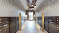 This thoughtfully designed mail room offers a refined blend of practicality and modern elegance. The space is lined on both sides with rows of dark metal mailboxes, providing ample secure mail and package storage for residents. Gold-toned pilasters punctuate the corridor at regular intervals, adding a warm, architectural rhythm that elevates the room's overall aesthetic. The ceiling draws the eye upward with a striking wood slat detail framed in rich brown tones, from which a series of warm Edison-style pendant lights hang at varying heights, casting a welcoming golden glow throughout the space. The walls feature a light stone-textured tile treatment that adds depth and texture without overwhelming the room. Underfoot, large-format light-toned flooring tiles laid in a clean pattern contribute to the bright, airy feel of the space. At the far end, a tall framed window allows natural light to filter in, creating a naturally illuminated focal point that enhances the sense of depth and openness. Subtle accent lighting at the base of the pilasters adds a finishing touch of sophistication. Overall, the space successfully transforms a utilitarian function into an experience that reflects quality craftsmanship and considered design.