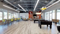 This single-floor community space is designed to accommodate a variety of social and recreational activities within one expansive, open-plan layout. The interior immediately draws attention with its striking exposed ceiling, which showcases an industrial framework of steel trusses, ductwork, and sleek linear pendant lighting — lending the space a modern, loft-like character.

The recreational zone is prominently anchored by a pool table and foosball table, providing engaging entertainment options. Adjacent to the game area is a comfortable lounge seating arrangement centered around a bold stone fireplace set against a vivid orange accent wall. Built-in shelving flanks the fireplace, and a wall-mounted television completes this inviting gathering spot.

A dedicated dining and café-style area occupies the opposite side of the space, outfitted with high-top bar tables and vibrant yellow seating. Large windows throughout allow generous amounts of natural light to flood the interior, enhancing the open and energetic atmosphere. Light wood-toned flooring runs throughout, tying the various zones together cohesively.

Overall, the space balances functionality with a lively, welcoming ambiance, making it well-suited for both casual socializing and organized group activities.