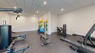 This fitness center offers a thoughtfully organized exercise space spread across a single floor. The room features a clean, modern aesthetic with crisp white walls, a suspended tile ceiling with recessed lighting, and durable dark rubberized flooring throughout — ideal for high-traffic athletic use.

Large windows along one wall allow natural light to flood the main exercise area, creating a bright and energizing environment. A wall-mounted split air conditioning unit ensures comfort during workouts year-round.

The space is well-appointed with a diverse selection of equipment. A multi-station cable and weight machine serves as a central fixture, complemented by a dedicated dumbbell rack stocked with a full range of free weights. Cardio equipment, including a stationary bike and additional aerobic machines, lines the windowed wall. A large wall mirror enhances the sense of space and supports form-checking during training.

A secondary area provides additional functional training space, housing a variety of stability and medicine balls on a tall multi-tier rack, along with adjustable benches and isolated resistance machines. This section offers ample open floor space for stretching or bodyweight exercises.

The overall layout is practical and well-organized, catering to a broad range of fitness routines from strength training to cardio and functional movement.