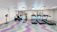 This fitness center at 5300 Eubank Blvd NE in Albuquerque, New Mexico, offers a well-equipped, single-floor exercise facility designed to support a well-rounded workout routine. The space is immediately distinguished by its bold chevron-patterned flooring in shades of purple, teal, green, and gray, lending the room a vibrant, energetic atmosphere.

The cardio area is outfitted with multiple treadmills, an elliptical trainer, and both upright and recumbent stationary bikes. Large windows line one wall, providing natural light and direct views of an outdoor swimming pool surrounded by mature trees — creating a bright, motivating environment for cardio workouts. Overhead, flush-mounted rectangular lighting fixtures ensure the space remains well-lit throughout.

A dedicated strength and functional training zone features a rack of dumbbells, medicine balls, kettlebells, and stability balls, along with wall-mounted workout charts and a wall-mounted flat-screen television for added convenience. The white walls keep the space feeling clean and open, while glass-paneled entry doors connect the gym to adjacent areas. A standing fan adds to the practical amenities available to users.

Overall, this fitness center combines functional design with lively aesthetics, offering a comfortable and fully equipped environment for cardio, strength, and functional training.